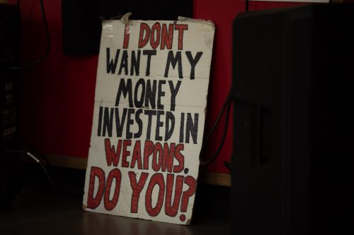 A white cardboard with 'I don't want my money invested in weapons. Do you?' written in caps. The 'I don't' and 'Weapons. Do you?' is painted in red with a black outline, whilst the rest is painted black caps.