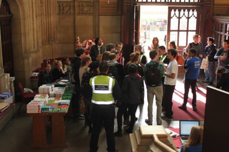 group of students standing in a grand university hall. Security guard in hi-vis jacket clearly visible standing by them.