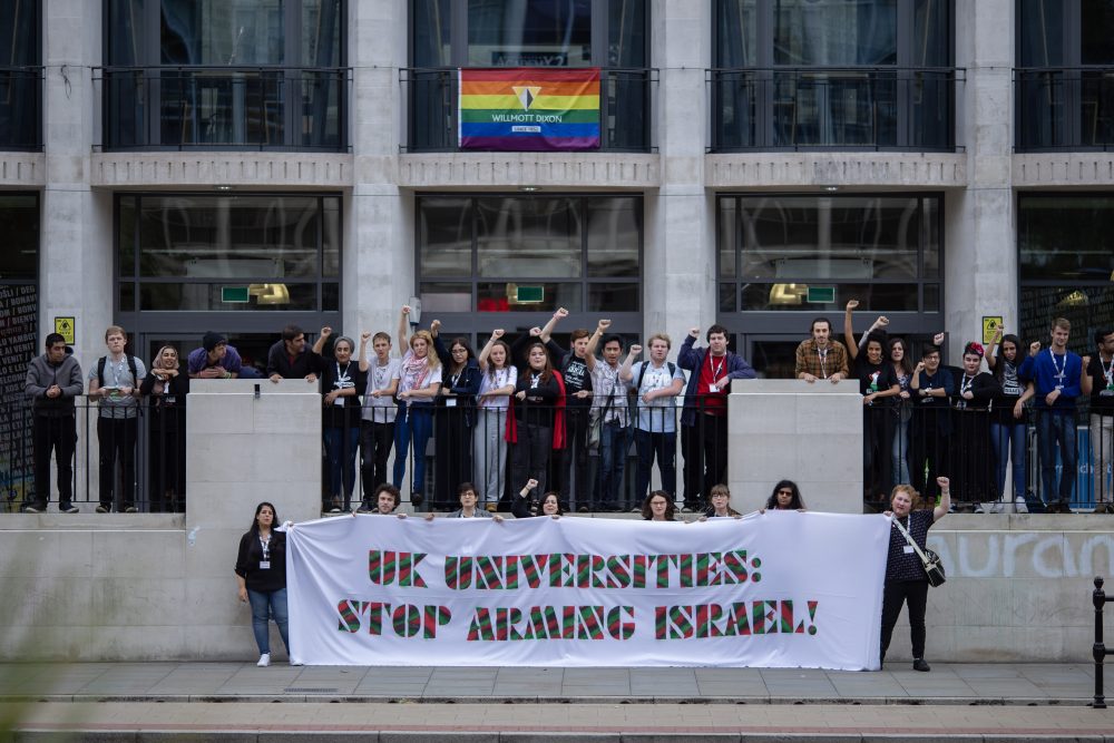 students stood outside the Manchester University Student Union building, many with fists up in the air. Two students on lower level/ground (in front of those standing on the platform above, behind a railing) hold a huge banner with the words 'UK UNIVERSITIES: STOP ARMING ISRAEL' in large multi-coloured caps. 