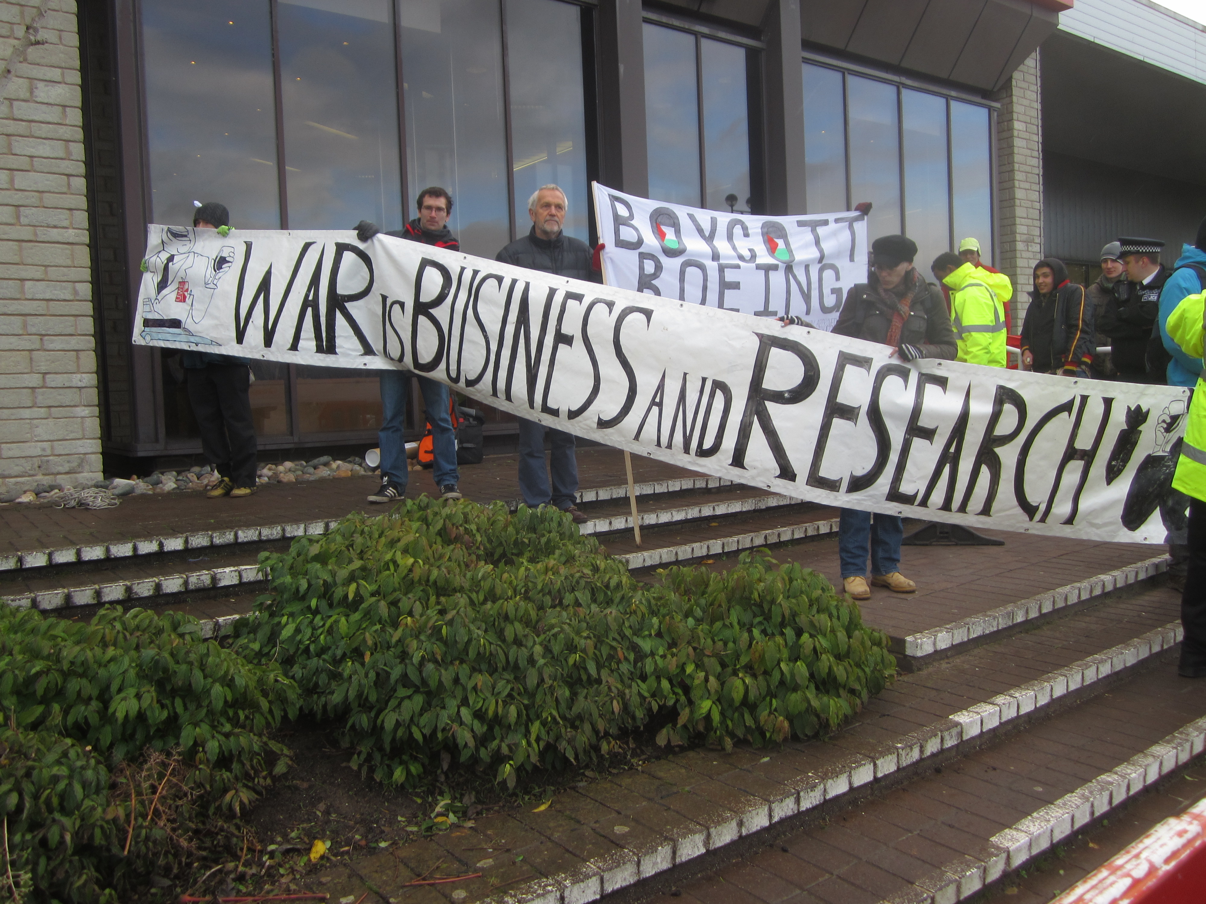 several protesters hold a banner stating that 'War is Business and Research'