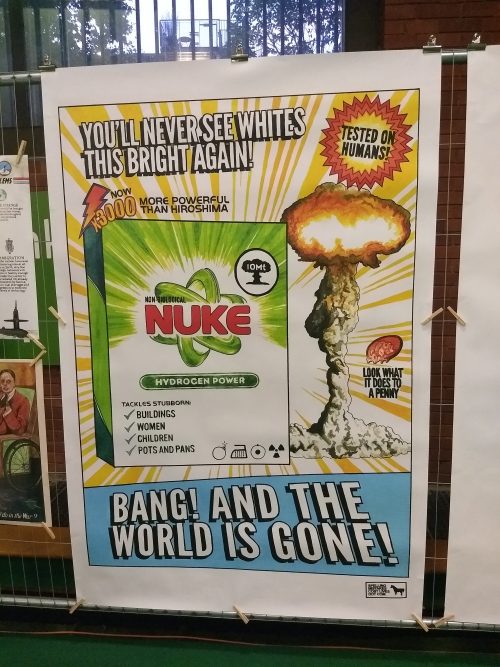 Poster with a box of detergent at the centre of the poster with thr word 'Nuke' in red caps on the box. Underneath 'Nuke' on the box, there is a list with 'Tackles Stubborn: Buildings, Women, Children, Pots and Pans'. Behind the box of detergent is an explosion with a 'tested on humans' sticker above. Next to it in big white caps is 'You'll Never see Whites This Bright Again!'. On top of the detergent box, in small black caps, it reads 'more powerful than hiroshima', with a 'now x3000' and lightining sign next to it. Beneath the box, there is 'Bang! And The World Is Gone!' in big white caps on a light blue background. The 