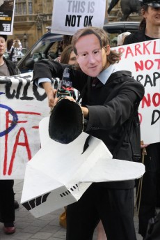 Protester with a David Cameron mask aims a fake gun at the camera