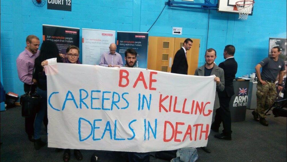 Three students hold banner outside BAE careers fair stall reading "BAE Careers in killing deals in death"