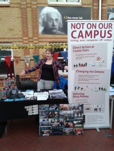 White women stands behind a stall covered in CAAT leaflets, with bunting and hazard tape behind her.