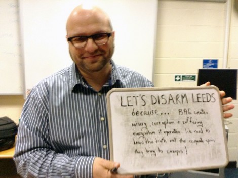 Andrew Feinstein holds board reading "Let's Disarm Leeds because... BAE creates misery, corruption and suffering everywhere it operates. We need to know this truth not the corporate spin they bring to campus!"